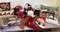 Full view of various Christmas pillows, throws, stuffed Santa and snowman decor, stockings, and fabric reindeer arranged on table against white wall