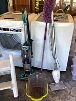 Shark Duo vacuum, purple duster, white electric mop with bucket, and two step stools in front of washer and dryer units.