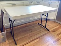 Side view of the folding table showing white plastic top and black metal folding legs on wood floor.