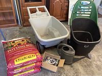 Full view of the wheelbarrows, watering can, three bags of potting soil, and box with disk lights on concrete floor in a garage.