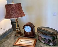 Photo showing brass table lamp with red paisley embroidered shade, wooden mantle clock, painted hat box and small framed print.