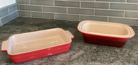 Two red Le Creuset baking dishes on countertop, one rectangular bread pan and one smaller casserole dish, both with cream interior.