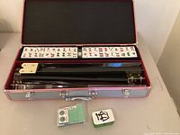 Open metal carrying case with a double row of Mahjong tiles, tile racks inside, small dice box, and green scoring sticks separated outside the case