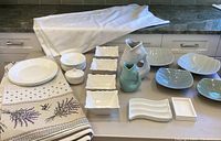Lot overview of white ceramic dishes, fish-shaped pitchers, and folded linens displayed on a countertop.