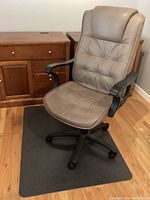 Office chair in taupe faux leather with padded seat and back, black armrests, and five-wheel base placed on dark grey floor mat in front of wooden cabinet.