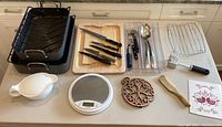 Full view of all items including two roasting pans with racks, wooden cutting board, cutlery set, ceramic gravy boat, digital scale, trivet, wooden spoon, and vegetable peeler.