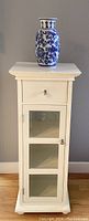 Front view of the white narrow cabinet with drawer and beveled glass door, blue and white porcelain vase on top.