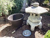 Front view of concrete lantern, plastic planter with coiled hose, and decorative metal plaque on ground