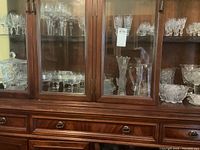 Display of various crystal and cut glassware items inside a wooden china cabinet. Includes stemmed glasses, vases, bowls, and a pitcher, showing clear intricate cut patterns.