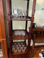 Three-shelf wooden stand holding several crystal glasses: top shelf with 8 tall wine and 2 stemmed wine glasses, middle and bottom shelf with various cocktail and juice glasses.
