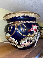 Front and side view of a dark blue ceramic floral planter with gold bows and leaf decorations, showing detailed hand-painted pink and white flowers and a scalloped gold rim.