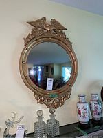Round gold framed mirror with eagle carved on top, wall mounted above two crystal decanters and ceramic vases.