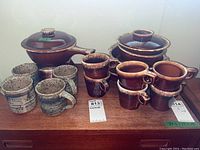 All items shown together on a wooden surface: six mugs, one casserole pot with lid, one covered soup bowl.