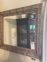 Frontal photo showing the entire ornate framed mirror leaning against a wall with a china cabinet reflection visible in the glass.