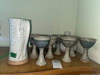 Pitcher, eight goblets, and lidded pot displayed on wooden surface with white walls background