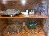 Shelf with multiple clear glass serving trays, clear glass pitcher, Avon perfume bottle with stopper, and three white ceramic duck figurines.