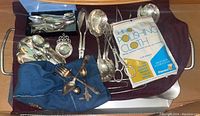 Silverplate, sterling spoons, various serving pieces including ladles, forks, and spoons grouped on a purple cloth with polishing cloth visible.