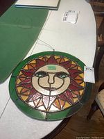 Photo of the circular stained glass sun catcher featuring a sun face with multicolor pattern and hanging wire, on a white table