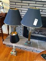 Three table lamps are shown together on a wooden floor in front of a brick fireplace. Two tall lamps have glass bases and brass stems with black shades, one small lamp with brass base and black shade is also visible.