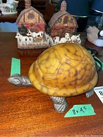 Photo of amber glass turtle lamp from side showing amber shell and metal base, with two red barn farm figurines with cows in background.