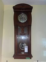 Full view of vintage wooden pendulum wall clock mounted on wall with visible pendulum and key.