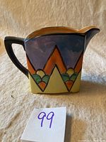Side angle of rectangular ceramic pitcher showing geometric orange and purple zigzag and green floral design on yellow background, black handle, and spout.