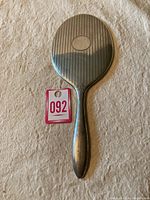 Photo of entire back of the oval hand mirror showing striped silver design and central oval medallion, alongside auction lot number tag