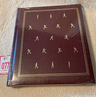 Front cover of The Incredible Machine book in maroon leather-like hardcover with gold embossed illustration of running human figures.
