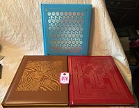 Three hardcover coffee table books: one blue with gold shell embossing, one brown with gold tiger design, one red with gold geometric embossing.