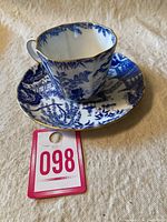 Demitasse teacup with handle, Blue Mikado chinoiserie pattern, and matching saucer shown on cloth background, with auction tag number 098 in the image.