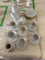 Bohemia Fine China Helene coffee set arranged on quilted surface including coffee pot, creamer, sugar bowl, cups and saucers, dessert plates. One cup visibly chipped.