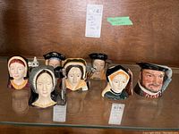 Front view of seven Royal Doulton Toby character mugs representing Henry VIII and his wives, arranged in a row on a glass shelf.