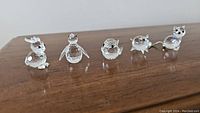 Front angle of all five clear crystal animal figurines (rabbit, penguin, bird, pig, cat) placed on wood surface