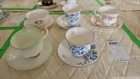 Photo showing all five teacups with matching saucers arranged on quilted surface, detailed floral and gold trim visible