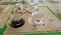 Four teacups and matching saucers displayed on fabric with visible floral designs and gold accents, showing different patterns including black and gold and white with pink roses.