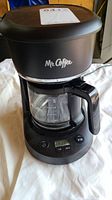 Front view of the Mr. Coffee coffeemaker showing brand logo, glass carafe with handle, control buttons, and power button.