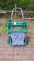 Front view of green plastic and metal garden tool dolly with fabric pocket holder and wheels on paving stones.