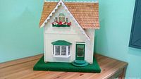 Front view of the handmade dollhouse showing white facade, green door, bay window with curtains, flower box, and green carpet base.