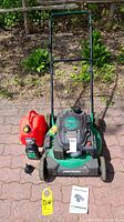 Front view of mower with accessories (gas can, oil, funnel, manuals) on paving stones