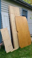 Various cut pieces of plywood sheets leaning against house, includes notched plywood, plain plywood, and smaller sheet with markings
