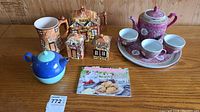 Photo showing all items on a wooden table: Brothers of England cottage teapot set, Chinese pink floral tea set on tray, blue and white nested teapot with cup, and recipe booklet.