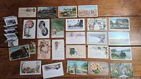Full lot of vintage postcards spread on wooden table, showing variety of scenes and artwork including floral designs, landscapes, buildings, and small folded accordion-style photo booklet.
