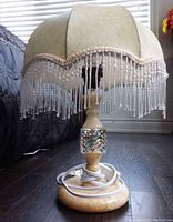 Full view of one marble base table lamp with scalloped beige fabric lampshade trimmed with beaded fringe and electrical cord at base.