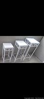 Set of three white metal nesting tables with scrollwork decoration, varying sizes, placed on concrete floor.