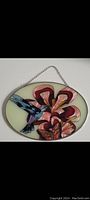 Oval stained glass artwork showing a blue and purple hummingbird feeding on red and pink flowers, pale green background, metal frame, hanging chain attached.