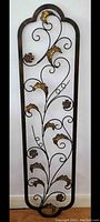Tall wrought metal wall trellis with scrolling vines and gold-accented leaves and flowers, standing against white wall to show full size and decorative details.