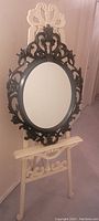 Full view of the oval mirror showing decorative dark frame with intricate detailing, placed against a light-colored easel stand.