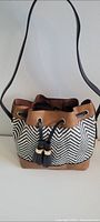Front and side view of Aldo bucket bag showing zigzag fabric body, brown trim, black drawstring with beads and adjustable black shoulder strap.