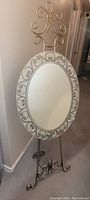 Oval mirror on decorative wrought iron stand, showing full front view including scroll and floral mosaic frame and curled iron base.