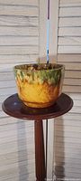 Green, yellow, and brown glazed decorative pot on a round wooden stand in front of white blinds.